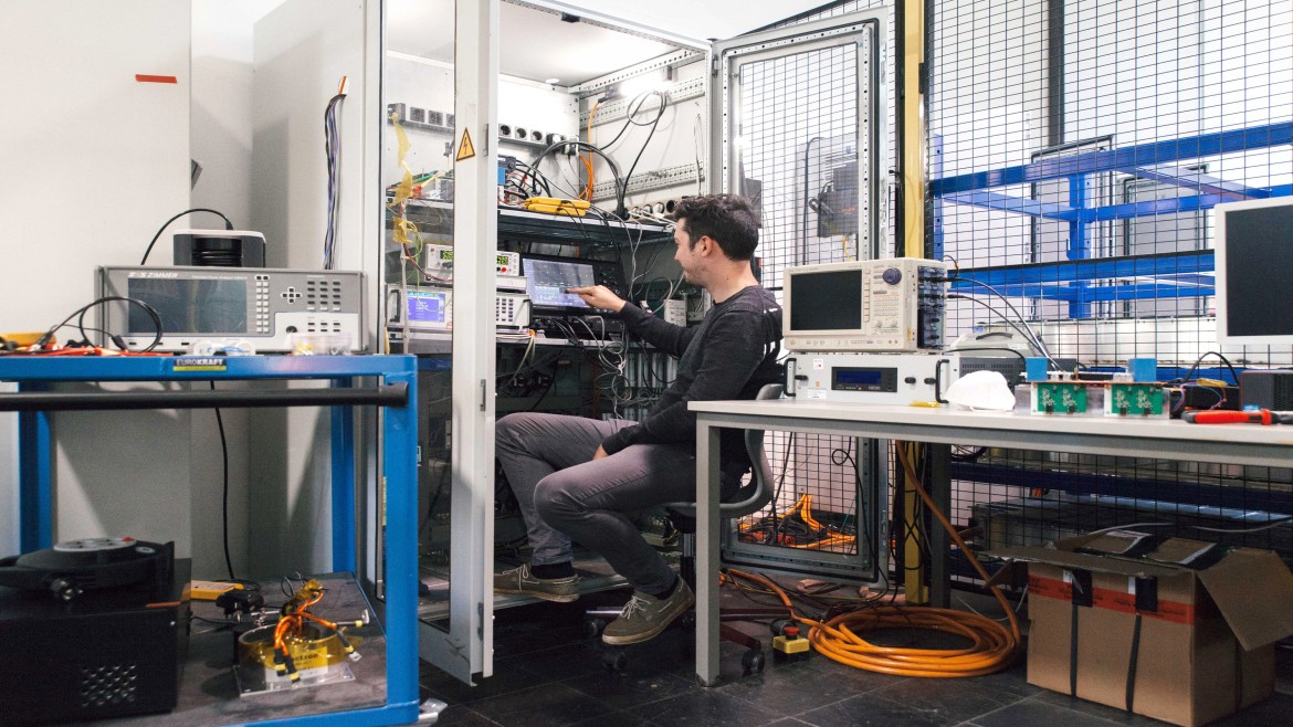 Ein Forscher sitzt auf einem Bürostuhl im Bildzentrum und bedient aufmerksam den Touchscreen eines Messmonitors, welcher in einen großen, geöffneten Elektronik-Schaltschrank integriert ist; der Schaltschrank ist mit diversen Kabeln, Stromversorgungen und Modulen bestückt; rechts im Vordergrund stehen ein digitales Oszilloskop sowie weitere Laborgeräte auf einem Tisch vor einer hohen Gittertrennwand, während auf einem blauen Rollwagen auf der linken Bildseite ein Zimmer-Leistungsanalysator platziert ist und die technische Laborumgebung vervollständigt.
