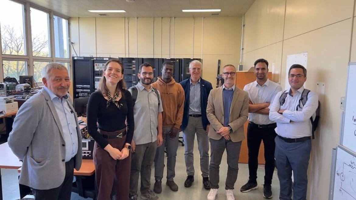 Ein Gruppenfoto in der Datei image_cf1888.jpg zeigt die bayerische Wissenschafts-Delegation mit acht Personen, die in einem hellen technischen Laborraum stehen und lächelnd in die Kamera blicken; die Gruppe setzt sich aus sieben Männern und einer Frau in geschäftlicher und legerer Kleidung zusammen; im Hintergrund des Raumes sind große Server- oder Elektronikschränke sowie Messplätze aufgebaut, während große Fenster auf der linken Seite Tageslicht hereinlassen.