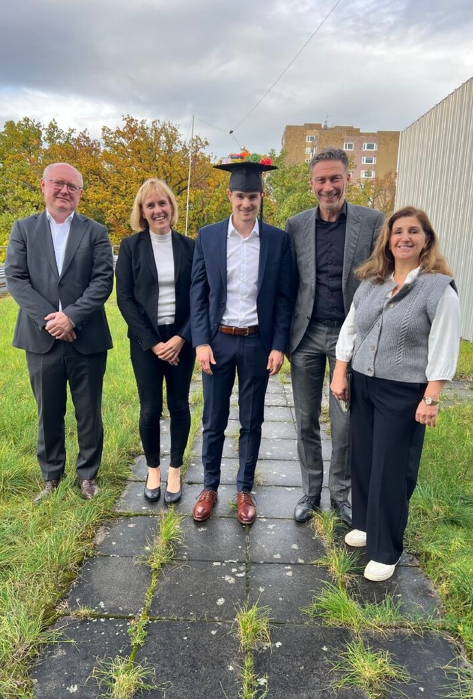 Ein Gruppenfoto im Freien zeigt fünf Personen nach einer erfolgreichen Promotion auf einer begrünten Dachfläche; in der Bildmitte steht der Promovierte Lukas Ranzinger im dunkelblauen Anzug und trägt stolz einen festlich geschmückten Doktorhut; er wird von vier lächelnden Kolleginnen und Kollegen in geschäftlicher Kleidung flankiert, die sich auf einem von Gras durchwachsenen Gehwegplatten-Pfad vor herbstlichen Bäumen und bewölktem Himmel präsentieren.